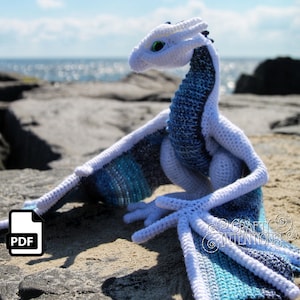 May include: A crocheted white and blue dragon with green eyes. The dragon is sitting on a rocky beach with the ocean in the background. The dragon has a long tail and wings. The dragon is a handmade amigurumi toy.
