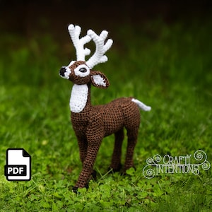Puede incluir: Una figura de ciervo de ganchillo con cuerpo marrón, cornamenta blanca y pecho blanco. El ciervo tiene ojos negros y está sobre hierba verde. Un icono PDF está en la esquina inferior izquierda y el texto "Crafty Intentions" está en la esquina inferior derecha.