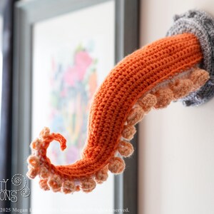 May include: A crocheted octopus tentacle wall hanging. The tentacle is orange with light orange suckers and emerges from a gray, circular wall mount. The piece is handmade and adds a whimsical touch to any space.