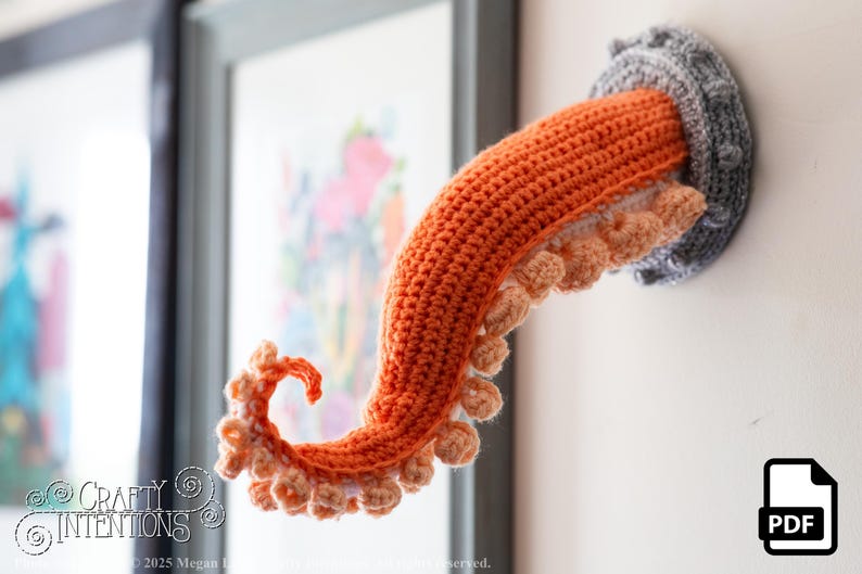 May include: A crocheted octopus tentacle wall hanging. The tentacle is orange with light orange suckers and emerges from a gray, circular wall mount. The piece is handmade and adds a whimsical touch to any space.