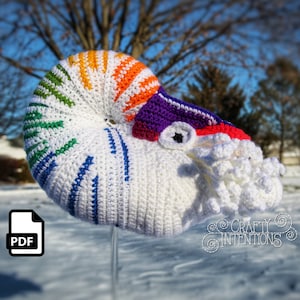 May include: A crocheted rainbow nautilus shell with a white crocheted body and a white crocheted tentacle. The shell is on a metal rod and is in the snow. The image includes the text "CRAFTY INTENTIONS".