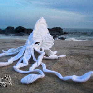 May include: White crocheted squid amigurumi toy on a sandy beach with ocean waves in the background. The squid has large, green eyes and long tentacles.