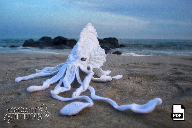 May include: White crocheted squid amigurumi toy on a sandy beach with ocean waves in the background. The squid has large, green eyes and long tentacles.