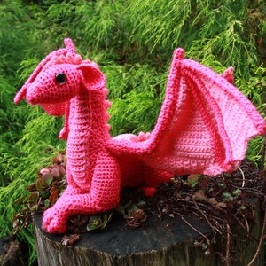 May include: A pink crocheted dragon with large wings. The dragon is sitting on a tree stump with green foliage in the background.
