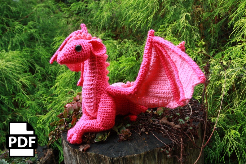 May include: A pink crocheted dragon with large wings. The dragon is sitting on a tree stump with green foliage in the background.