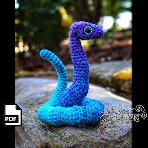 Könnte beinhalten: Ein gehäkeltes Schlangen-Amigurumi-Spielzeug mit einem blauen und violetten Farbverlauf. Die Schlange ist zu einer Spirale zusammengerollt, mit dem Kopf erhoben. Die Schlange hat ein schwarzes Auge mit einer blauen Pupille.