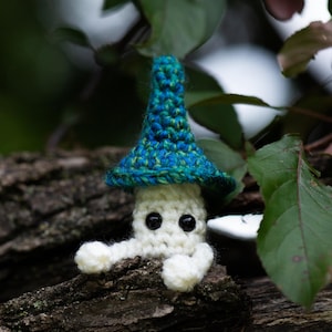 Puede incluir: Una figura de hongo hecha a mano con ganchillo, con cuerpo blanco, ojos negros y un sombrero azul y verde. El hongo est&aacute; posado sobre un trozo de madera, rodeado de hojas verdes. El texto "Crafty Intentions" y un icono PDF son visibles.
