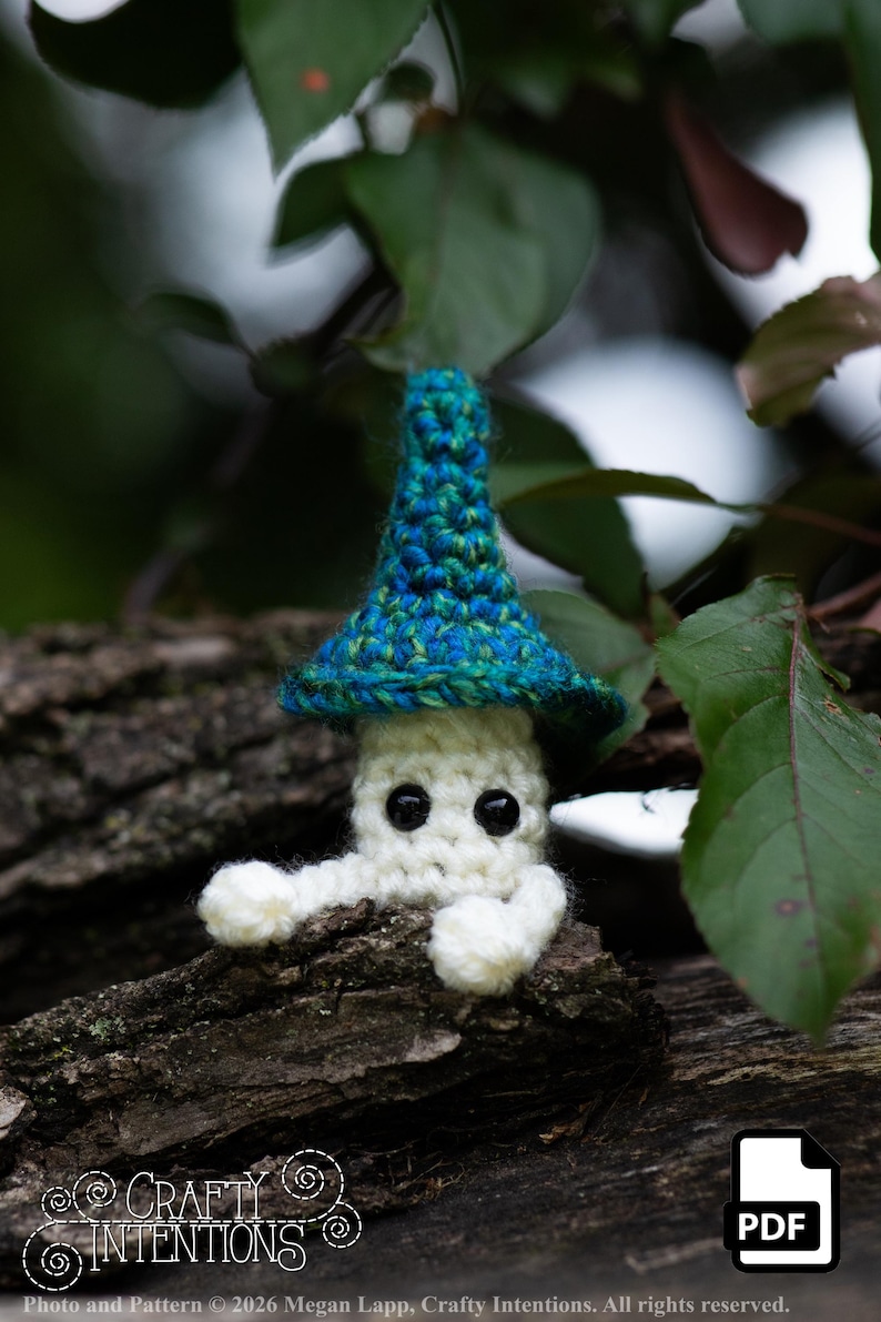 Puede incluir: Una figura de hongo hecha a mano con ganchillo, con cuerpo blanco, ojos negros y un sombrero azul y verde. El hongo est&aacute; posado sobre un trozo de madera, rodeado de hojas verdes. El texto "Crafty Intentions" y un icono PDF son visibles.