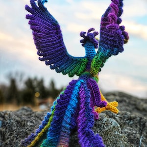 May include: A colorful crocheted phoenix with outstretched wings, standing on a rock. The phoenix is made with a rainbow of colors, including purple, blue, green, and yellow.