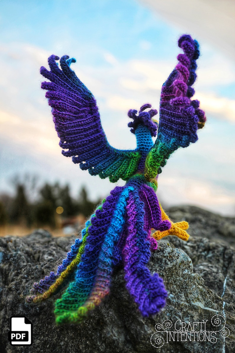 May include: A colorful crocheted phoenix with outstretched wings, standing on a rock. The phoenix is made with a rainbow of colors, including purple, blue, green, and yellow.