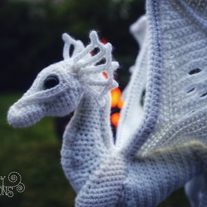 May include: White crocheted dragon with a large wing, a detailed head, and a black eye. The dragon is posed with its wing outstretched. The background is out of focus and appears to be green.