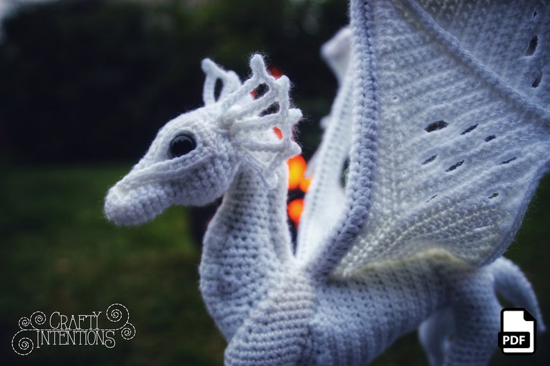 May include: White crocheted dragon with a large wing, a detailed head, and a black eye. The dragon is posed with its wing outstretched. The background is out of focus and appears to be green.