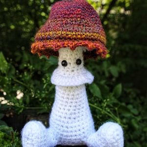 May include: A crocheted mushroom amigurumi toy with a white body and a red, orange, and purple cap. The toy has black eyes and is sitting on a wooden surface. The text "CRAFTY INTENTIONS" is visible on the image.