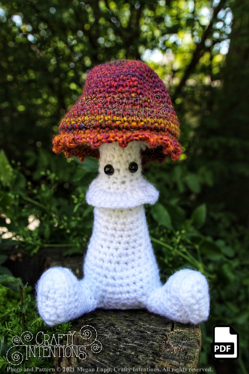 May include: A crocheted mushroom amigurumi toy with a white body and a red, orange, and purple cap. The toy has black eyes and is sitting on a wooden surface. The text "CRAFTY INTENTIONS" is visible on the image.