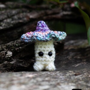 Puede incluir: Un hongo de ganchillo hecho a mano con un cuerpo blanco, ojos negros y un sombrero colorido. El sombrero presenta tonos de morado, azul y rosa. El hongo se encuentra sobre un fondo natural borroso. La imagen incluye el texto "Crafty Intentions" y un icono PDF.