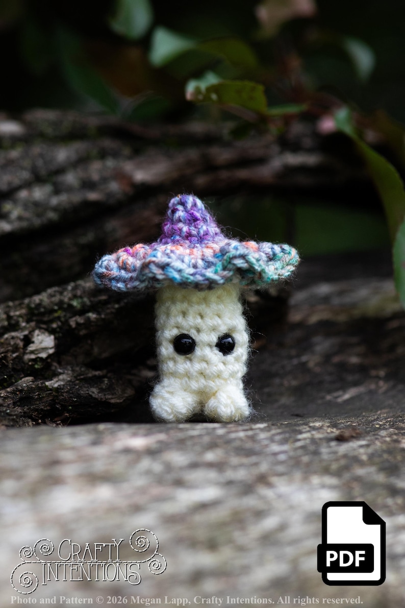 Puede incluir: Un hongo de ganchillo hecho a mano con un cuerpo blanco, ojos negros y un sombrero colorido. El sombrero presenta tonos de morado, azul y rosa. El hongo se encuentra sobre un fondo natural borroso. La imagen incluye el texto "Crafty Intentions" y un icono PDF.