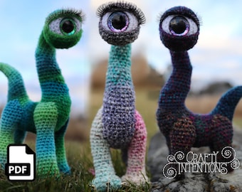Può includere: Tre giocattoli amigurumi all'uncinetto a forma di creatura dal collo lungo con un solo occhio grande. I giocattoli sono in combinazioni di colori verde, viola e blu. I giocattoli sono seduti su una superficie erbosa con una roccia sullo sfondo. L'immagine include un'icona PDF nell'angolo in basso a sinistra.