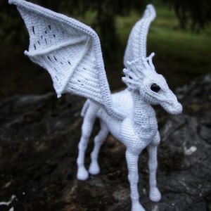 May include: White crocheted hippogriff with wings outstretched. The hippogriff is standing on a dark brown surface.