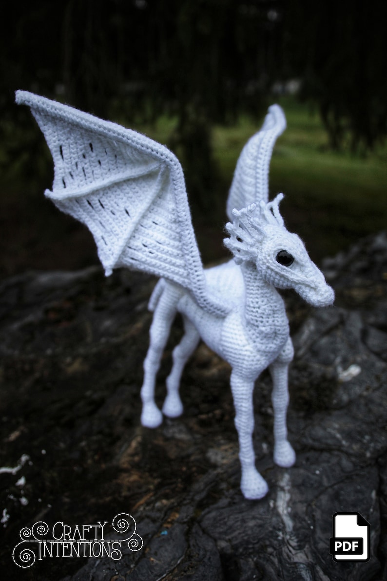 May include: White crocheted hippogriff with wings outstretched. The hippogriff is standing on a dark brown surface.