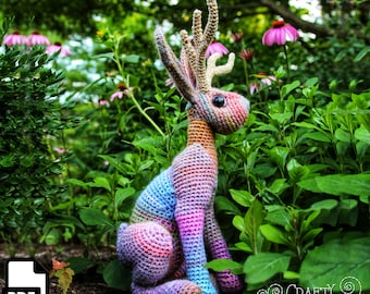 May include: A colorful crocheted rabbit with antlers, sitting in a garden setting. The rabbit is made with a variety of shades of pink, blue, and purple yarn. The antlers are made with a brown yarn. The rabbit is sitting on a bed of green leaves. The image includes the text "CRAFTY INTENTIONS" and a PDF icon.