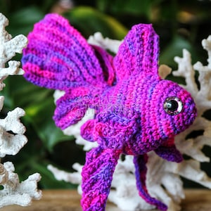 Puede incluir: Un pez betta de crochet morado y rosa con un ojo brillante. El pez est&aacute; sentado sobre una superficie de madera con coral blanco detr&aacute;s. La imagen incluye el texto "CRAFTY INTENTIONS" y "PDF".