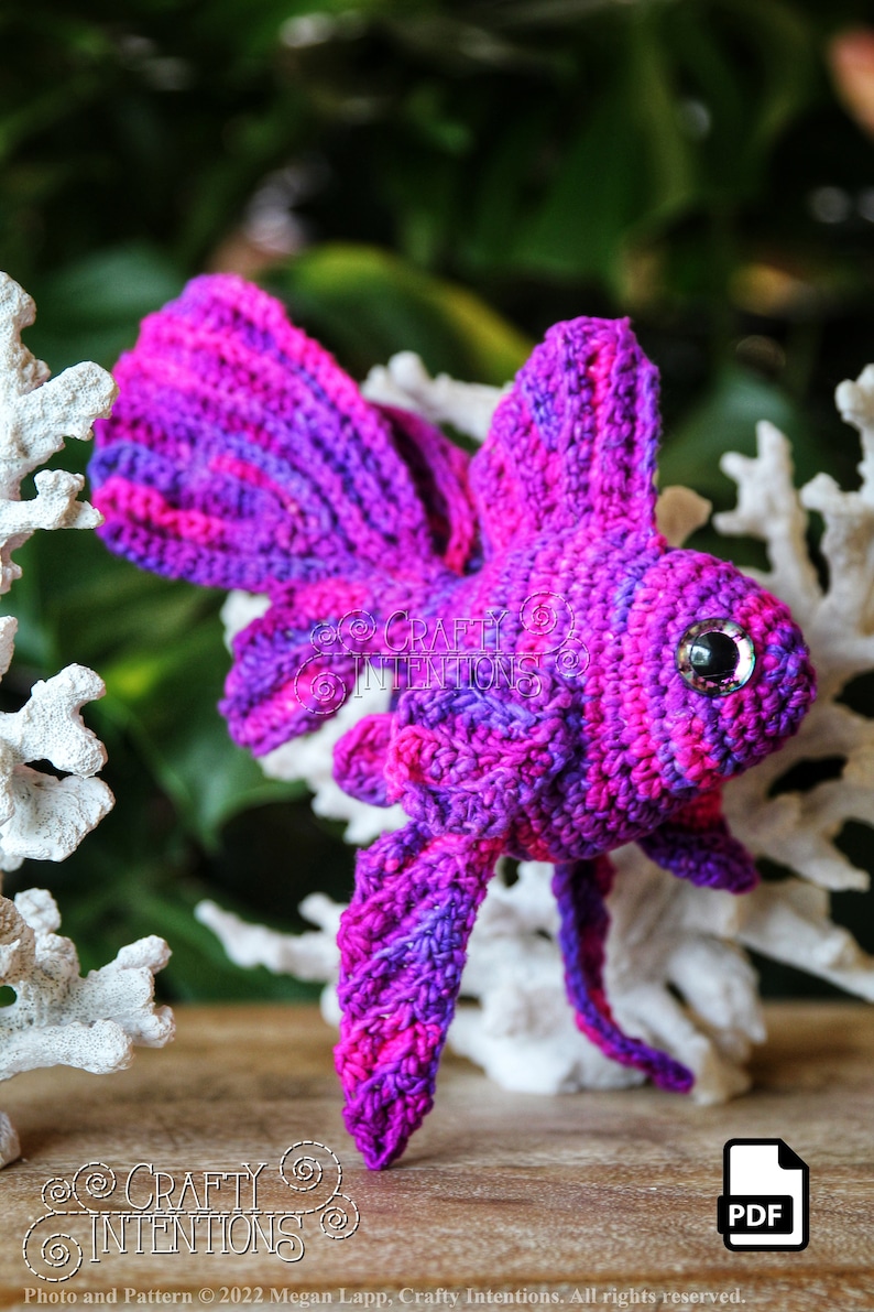Puede incluir: Un pez betta de crochet morado y rosa con un ojo brillante. El pez est&aacute; sentado sobre una superficie de madera con coral blanco detr&aacute;s. La imagen incluye el texto "CRAFTY INTENTIONS" y "PDF".