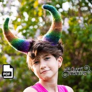 Peut inclure: Une personne portant un bandeau en crochet arc-en-ciel avec des cornes. Le bandeau est fait de fil de laine dans des tons de violet, vert, orange et jaune. La personne regarde la caméra et sourit.