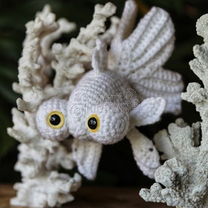 Puede incluir: Juguete amigurumi de pez dorado de ganchillo blanco con ojos amarillos, posado sobre un fondo de arrecife de coral blanco. El juguete es un art&iacute;culo artesanal hecho a mano y est&aacute; disponible como patr&oacute;n PDF descargable.