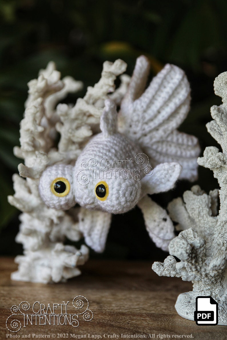 Puede incluir: Juguete amigurumi de pez dorado de ganchillo blanco con ojos amarillos, posado sobre un fondo de arrecife de coral blanco. El juguete es un art&iacute;culo artesanal hecho a mano y est&aacute; disponible como patr&oacute;n PDF descargable.