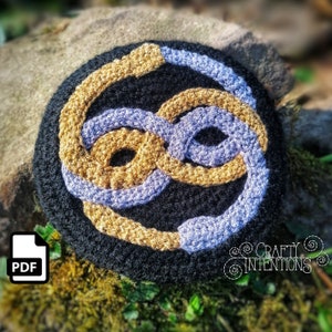 Könnte beinhalten: Gehäkeltes Unendlichkeitszeichen in goldener und lilafarbener Wolle auf schwarzem Hintergrund. Das Symbol ist ein beliebtes Design für Schmuck, Wohnaccessoires und andere Handarbeiten.