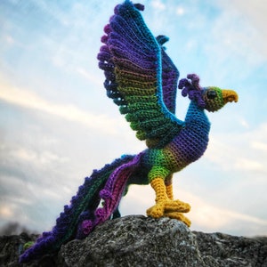 May include: A colorful crocheted phoenix with a purple, green, and yellow color scheme stands on a gray rock with a blue sky in the background. The phoenix has its wings spread out and its tail feathers are flowing behind it. The image includes the text "CRAFTY INTENTIONS" in the lower right corner.