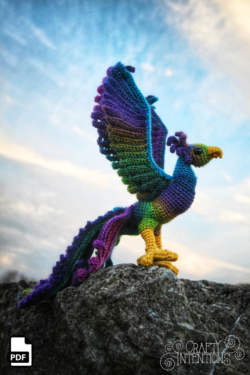 May include: A colorful crocheted phoenix with a purple, green, and yellow color scheme stands on a gray rock with a blue sky in the background. The phoenix has its wings spread out and its tail feathers are flowing behind it. The image includes the text "CRAFTY INTENTIONS" in the lower right corner.
