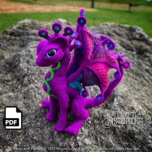 May include: A crocheted purple dragon with green accents and iridescent wings. The dragon has large blue eyes and is sitting on a rock.