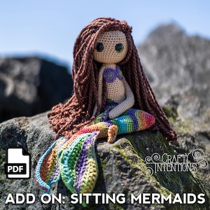 May include: A crocheted mermaid doll with brown hair and a rainbow tail sits on a rock. The doll has a beige body, a purple top, and large black eyes. The text "ADD ON: SITTING MERMAIDS" is at the bottom of the image.
