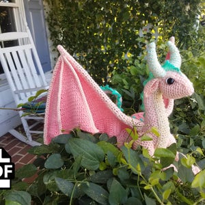 K&ouml;nnte beinhalten: Ein rosa geh&auml;kelter Drache mit gr&uuml;nen Akzenten und einem gro&szlig;en Fl&uuml;gel. Der Drache sitzt in einem Bett aus gr&uuml;nen Bl&auml;ttern.