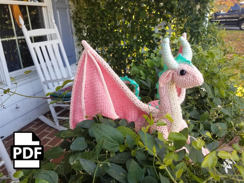 K&ouml;nnte beinhalten: Ein rosa geh&auml;kelter Drache mit gr&uuml;nen Akzenten und einem gro&szlig;en Fl&uuml;gel. Der Drache sitzt in einem Bett aus gr&uuml;nen Bl&auml;ttern.