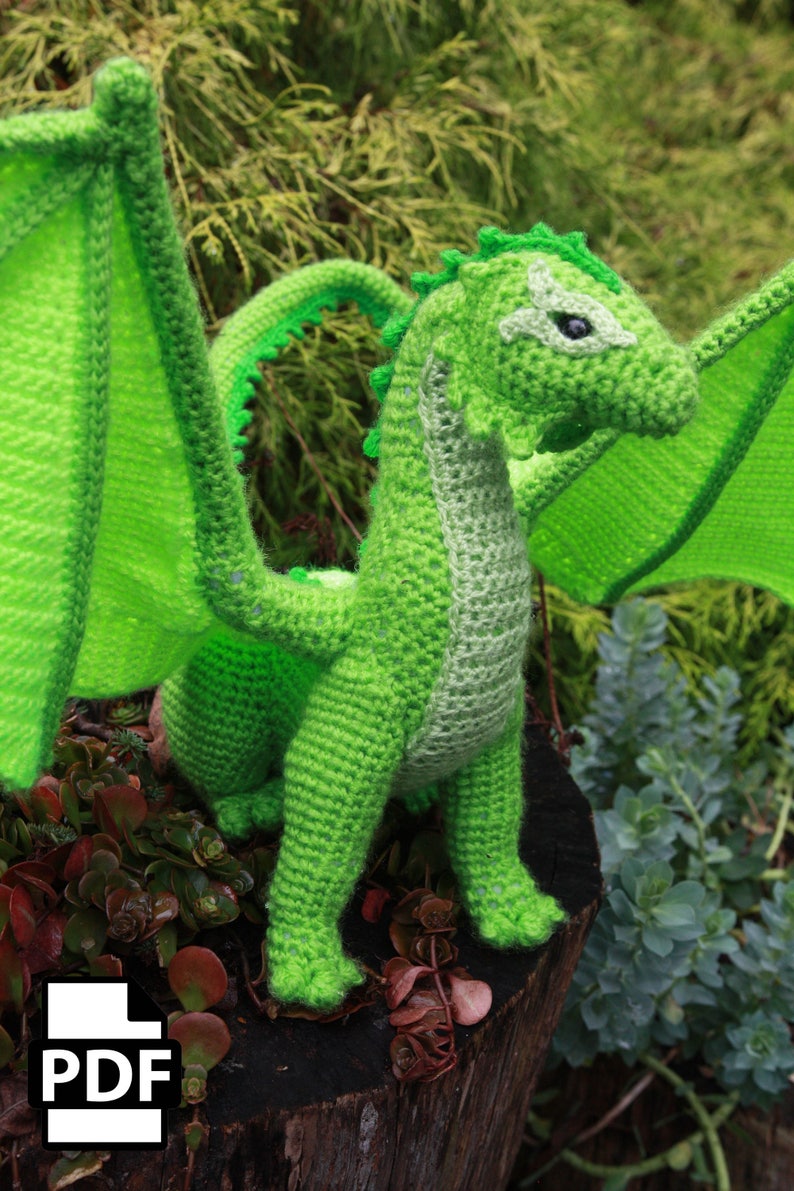 K&ouml;nnte beinhalten: Ein gr&uuml;ner geh&auml;kelter Drache mit Fl&uuml;geln, der auf einem Baumstumpf steht. Der Drache hat ein detailliertes Gesicht und einen strukturierten K&ouml;rper.