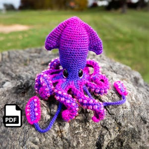 May include: A crocheted squid amigurumi toy with a pink and purple ombre color scheme. The squid has large, black eyes and a textured body. The tentacles are decorated with small, pink beads.