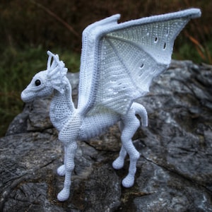 May include: White crocheted dragon with wings outstretched, standing on a gray rock. The dragon has a detailed face and legs. The wings are made with a lacy stitch.