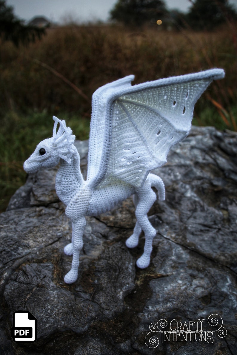 May include: White crocheted dragon with wings outstretched, standing on a gray rock. The dragon has a detailed face and legs. The wings are made with a lacy stitch.