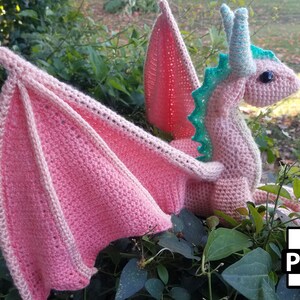 May include: A pink crocheted dragon with green accents and large black eyes. The dragon is sitting on a bed of green leaves. The wings are spread out behind the dragon. The image includes the word "PDF" in the lower right corner.