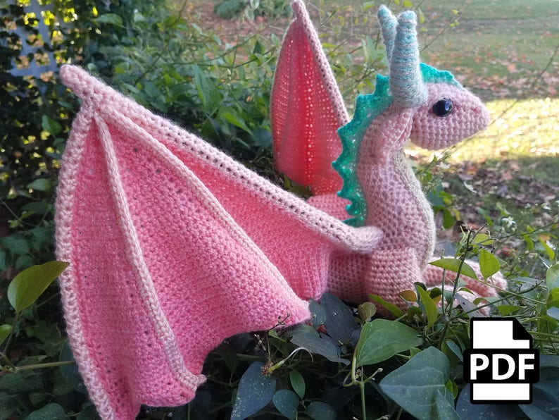 May include: A pink crocheted dragon with green accents and large black eyes. The dragon is sitting on a bed of green leaves. The wings are spread out behind the dragon. The image includes the word "PDF" in the lower right corner.