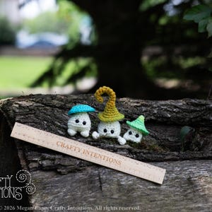 Puede incluir: Tres figuras de setas de ganchillo con cuerpos blancos y gorras coloridas en azul, amarillo y verde. Se exhiben sobre una superficie de madera desgastada con una regla que dice "Crafty Intentions". El fondo est&aacute; borroso, lo que sugiere un entorno al aire libre.