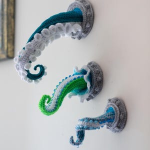 May include: Three crochet octopus tentacles in shades of blue, green, and white emerge from gray circular wall mounts. The tentacles have textured details and are mounted on a white wall. The Crafty Intentions logo is in the bottom left.