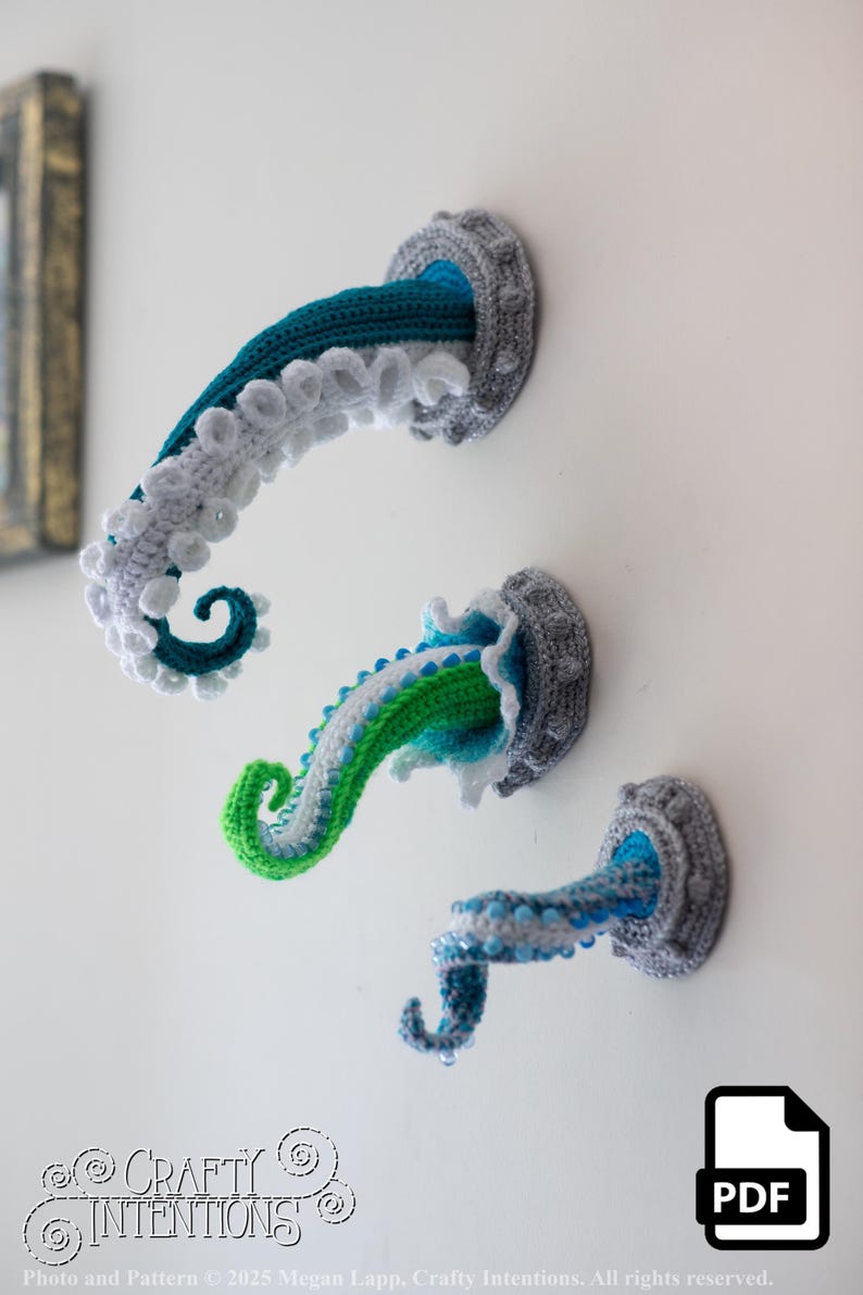 May include: Three crochet octopus tentacles in shades of blue, green, and white emerge from gray circular wall mounts. The tentacles have textured details and are mounted on a white wall. The Crafty Intentions logo is in the bottom left.