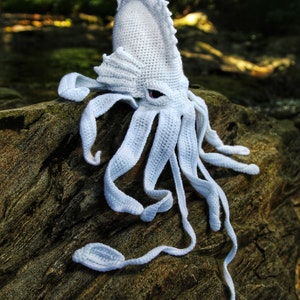 May include: White crocheted squid amigurumi pattern. The pattern is available as a PDF download. The squid has a large head with a pointed fin and eight tentacles.