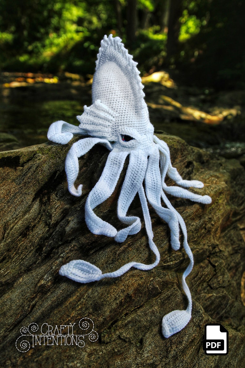 May include: White crocheted squid amigurumi pattern. The pattern is available as a PDF download. The squid has a large head with a pointed fin and eight tentacles.