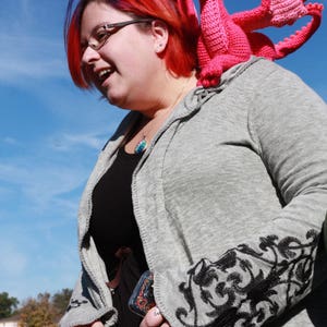 May include: A woman with red hair is wearing a gray cardigan with black floral embroidery. She is holding a pink crocheted dragon on her shoulder.