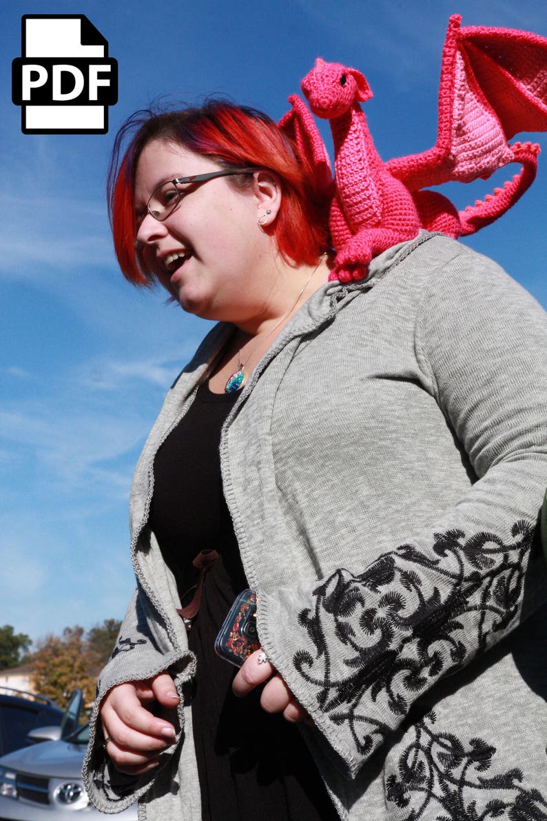 May include: A woman with red hair is wearing a gray cardigan with black floral embroidery. She is holding a pink crocheted dragon on her shoulder.