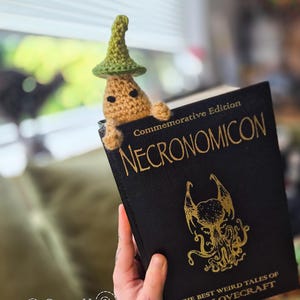Puede incluir: Un marcap&aacute;ginas de ganchillo con forma de hongo caprichoso con un sombrero verde, asom&aacute;ndose de un libro negro titulado "NECRONOMICON". El libro tiene letras doradas y una ilustraci&oacute;n dorada. Tambi&eacute;n es visible el texto "THE BEST WEIRD TALES OF H. P. LOVECRAFT".